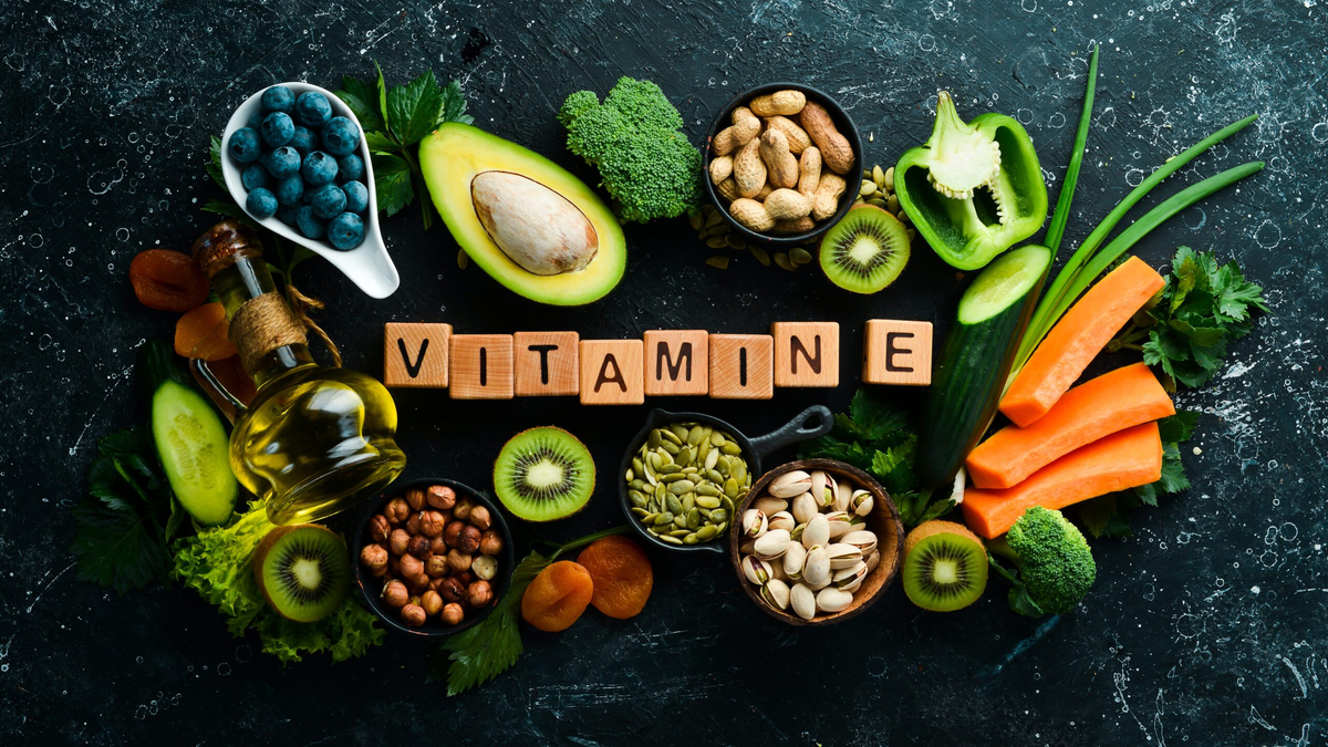 Wooden blocks with letters spelling out vitamin E on a marble table with bowls of nuts and fruits around the lettering